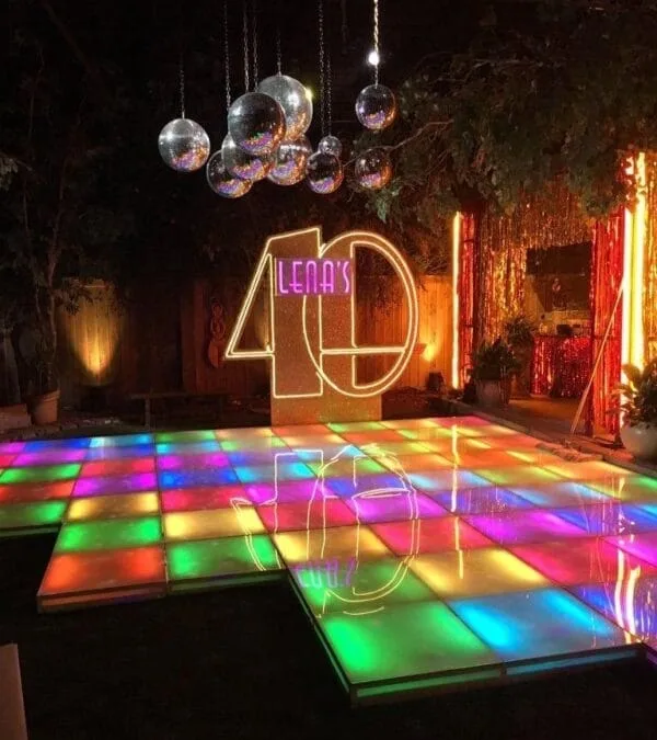 The LK - CH01 Rainbow LED Dance Floor was on display at the 40th birthday celebration. The dance floor was lit up with colorful lights, creating a festive atmosphere. Several disco balls were hung above the dance floor, reflecting light throughout the space. Decorative elements such as string lights and green plants added to the celebratory atmosphere.