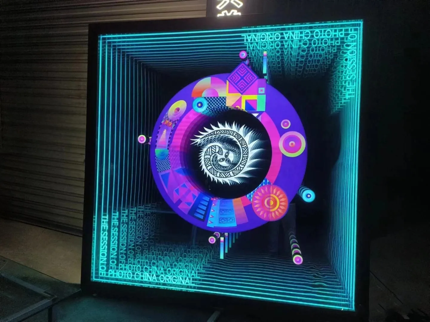Indywidualny ekran LED od Lucky Light A customized LED display featuring a vibrant and intricate graphic design with a circular central motif surrounded by colorful shapes and text. The display is set against a dark background, highlighting the bright and detailed visuals. The text on the display includes "THE SESSION" and other phrases, contributing to the overall artistic and technological aesthetic.